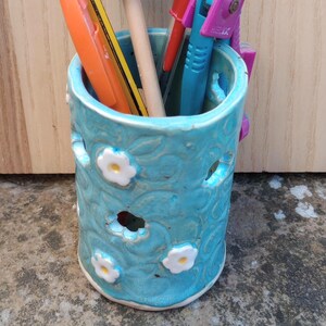 May include: A cylindrical turquoise ceramic pencil holder with embossed floral patterns and white flower accents, containing an orange marker, a yellow pencil, a blue house-shaped topper, a red pen, and a purple drafting compass tool.