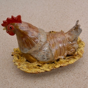 May include: A ceramic rooster figurine with a red comb and wattles, sitting on a yellow nest-like base.