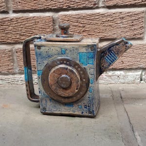 May include: A ceramic teapot with a unique, geometric design. The teapot features a square body with a circular element on the front, accented with blue and brown tones. The handle and spout are integrated into the design, creating a modern aesthetic.