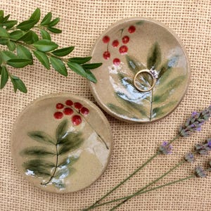May include: Two handmade ceramic ring dishes. Each dish is round and beige, with a painted design of green leaves and red berries. One dish holds a gold ring with a diamond. The dishes are on a hessian surface.