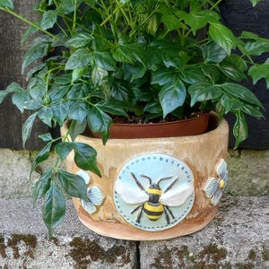 May include: A cylindrical ceramic planter with a tan, wood-grain finish featuring a central circular relief of a yellow and black bumblebee on a light blue background, flanked by two small, light blue and white sculpted flower blossoms.