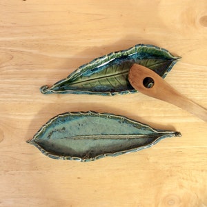May include: Two ceramic leaf-shaped spoon rests with a wooden spoon. The spoon rests are glazed in shades of green and blue, with a textured leaf vein design. The wooden spoon has a circular hole in the handle.