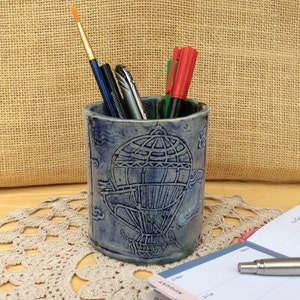 May include: Blue ceramic pencil holder featuring a hot air balloon design. The holder is filled with pens and pencils.