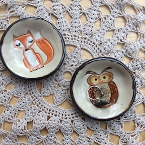 May include: Two ceramic ring dishes, one with a fox design and the other with an owl design. Both dishes are brown with a white glaze and are sitting on a crocheted doily.