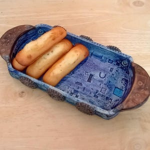 May include: A blue ceramic bread basket with a circuit board design holds three golden-brown breadsticks. The basket has brown accents on the ends and sides. The breadsticks are fresh and ready to eat.