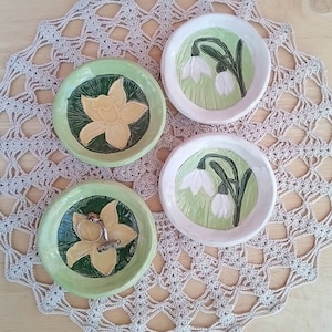 May include: Four ceramic trinket dishes with floral designs. Two dishes feature white snowdrop flowers on a green background. The other two dishes feature yellow daffodil flowers on a green background.