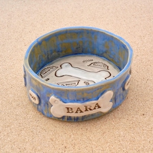 May include: A ceramic dog bowl with a blue and gold rim. The interior is white with a bone design and the name "BARA" on the front. The bowl has the words "Woof" and "Love me" on the sides. A pet accessory.