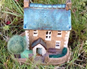 May include: A miniature ceramic house with a blue-green roof, brown brick walls, and white windows. The house has a small front porch and a green tree in the yard. The house is sitting on a brown base with a green grass border.