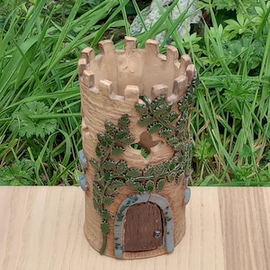 May include: A cylindrical, beige ceramic castle-shaped planter with a crenellated top. The planter features a small arched door, green ivy detailing, and blue accents. The planter is set on a wooden surface with green grass in the background.
