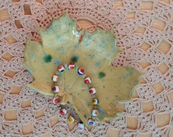 May include: A colorful glass bead bracelet with red, white, blue, and green beads is displayed on a ceramic leaf-shaped dish. The dish is resting on a crocheted doily.