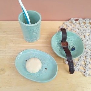 May include: A set of three light blue ceramic bathroom accessories with a mandala design. The set includes a toothbrush holder, a soap dish, and a small dish for jewellery or other small items.