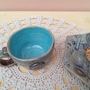 May include: A blue ceramic mug with a brown handle and a decorative design. The mug is sitting on a white crocheted doily. A ceramic plate with a brown and white design is in the background.