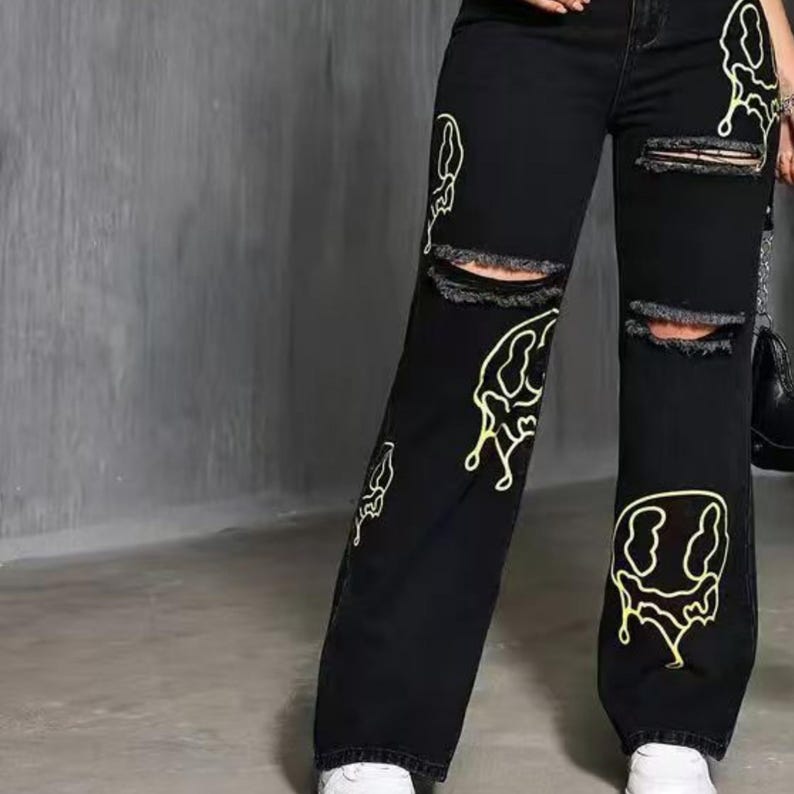 Pu&ograve; includere: Jeans neri in denim con ginocchia strappate e gamba larga. I jeans presentano un design a faccina sorridente gialla e altri disegni astratti gialli. La modella indossa scarpe da ginnastica bianche.