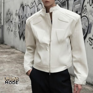 Puede incluir: Una chaqueta blanca con cuello alzado, cremallera frontal completa y detalles acolchados triangulares en relieve en el pecho y los hombros. La chaqueta presenta mangas largas y un corte estructurado, combinada con pantalones negros.