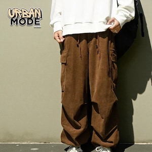 May include: Brown corduroy cargo pants with a drawstring waist and large side pockets. The pants are part of the Urban Mode collection, as indicated by the logo in the upper left corner. The pants are worn with a white sweatshirt.