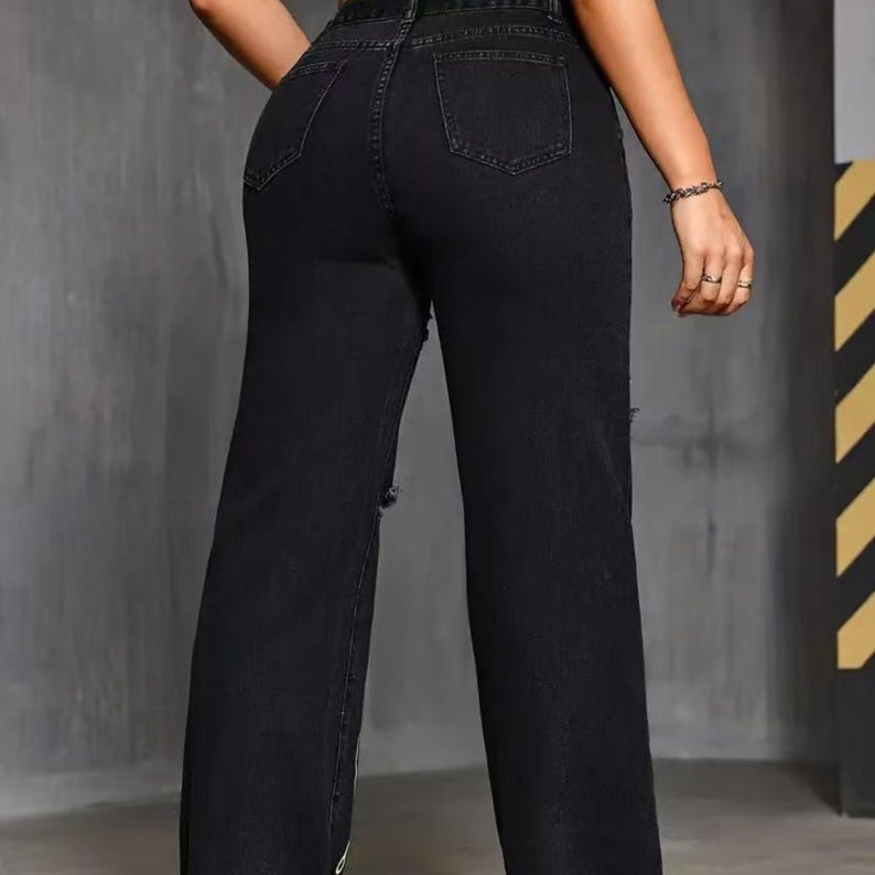 Pu&ograve; includere: Jeans neri a vita alta e gamba dritta. I jeans presentano dettagli strappati sulle ginocchia e tasche sul retro. La modella indossa un braccialetto e anelli.