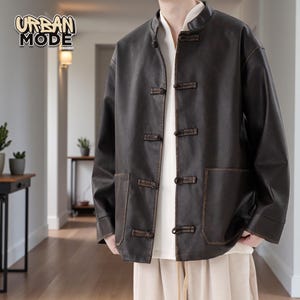 Puede incluir: Chaqueta marrón de cuero sintético con cuello mandarín y cierres de palanca de madera. La chaqueta tiene dos bolsillos delanteros y el logo "URBAN MODE" en la esquina superior izquierda. Se usa sobre una camisa blanca.