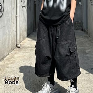 May include: Black denim cargo shorts featuring a loose, wide-leg fit, two large side cargo pockets with flaps, and two front pockets. The shorts include a black fabric tag attached to the waistband and are paired with black leggings and white sneakers.