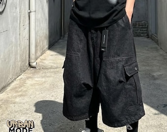 May include: Black denim cargo shorts featuring a loose, wide-leg fit, two large side cargo pockets with flaps, and two front pockets. The shorts include a black fabric tag attached to the waistband and are paired with black leggings and white sneakers.