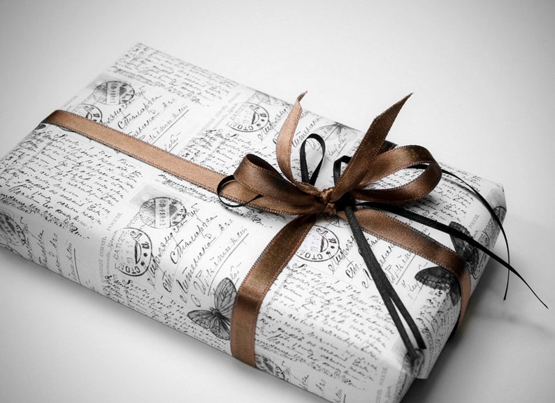 May include: A wrapped gift with vintage-style paper featuring script and butterfly illustrations. The gift is tied with a brown satin ribbon and a black ribbon, creating a decorative bow. The overall aesthetic is elegant and classic.