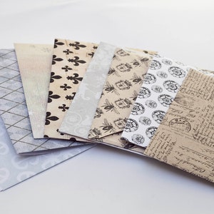 May include: A collection of decorative paper envelopes with various patterns. Designs include fleur-de-lis, bees, and vintage script. The envelopes are in shades of beige, gray, and white, suitable for crafts or stationery.
