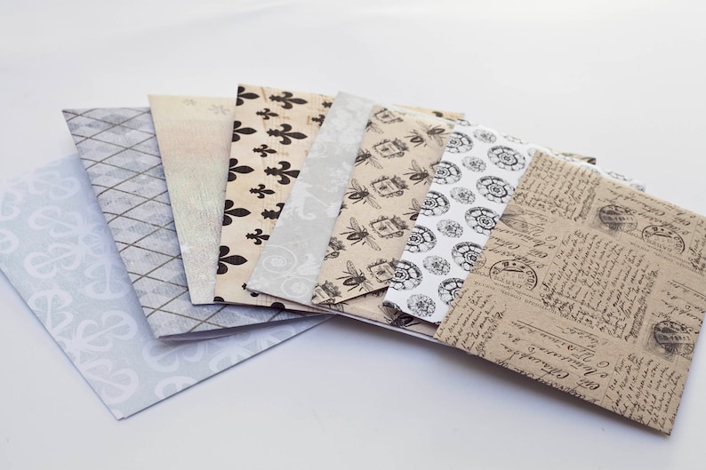 May include: A collection of decorative paper envelopes with various patterns. Designs include fleur-de-lis, bees, and vintage script. The envelopes are in shades of beige, gray, and white, suitable for crafts or stationery.