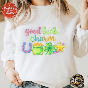 Puede incluir: Sudadera blanca con la frase "good luck charm" en colores vibrantes. El diseño incluye una herradura, una olla de oro, un trébol de cuatro hojas y una estrella.