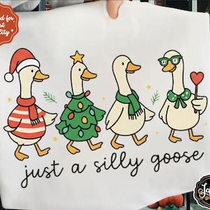 May include: A white fabric with a whimsical design of four geese. Each goose is dressed in festive outfits: a Santa hat and scarf, a Christmas tree, a green scarf, and glasses with a heart. The text reads "just a silly goose."