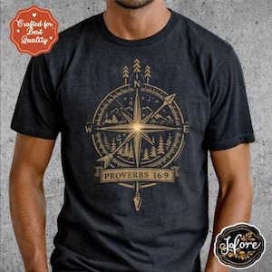 May include: Dark gray t-shirt featuring a gold compass design with mountains and trees. The design includes the text "PROVERBS 16:9". The shirt has a round neck and short sleeves.