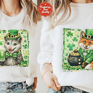 Puede incluir: Camiseta blanca de manga larga con un gráfico del Día de San Patricio. El gráfico presenta una zarigüeya de dibujos animados con un sombrero verde y pajarita, sosteniendo una cerveza verde. La otra camisa presenta un zorro con un pote de oro.