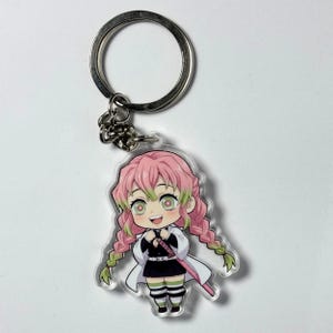 Könnte beinhalten: Ein Schlüsselanhänger mit einem Anime-Charakter im Chibi-Stil mit rosa Zöpfen, grünen Augen und einem weißen und schwarzen Outfit. Der Schlüsselanhänger hat einen silbernen Metallring und eine Kette.