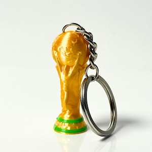 May include: A gold-colored keychain featuring a miniature trophy figure holding a globe aloft. The base of the trophy is accented with two horizontal green stripes. A silver-toned metal chain and a circular split ring are attached to the top.