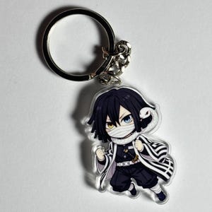 Könnte beinhalten: Ein klarer Acryl-Schlüsselanhänger mit einem Anime-Charakter im Chibi-Stil mit schwarzen Haaren, einem weißen Verband und einer Schlange. Die Figur trägt einen schwarz-weiß gestreiften Mantel und eine schwarze Hose. Der Schlüsselanhänger hat einen silbernen Metallring.