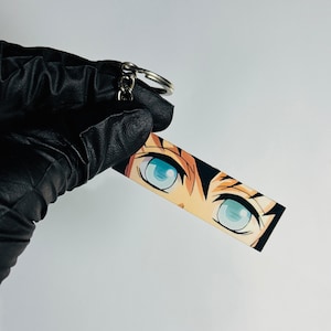 May include: A rectangular keychain with an anime-style illustration of a character's eyes. The eyes are bright blue, set against tan skin and black hair. The keychain has a silver keyring attached.