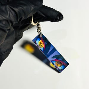 Peut inclure: Un porte-clés rectangulaire avec un motif d'œil de style anime aux accents bleus, jaunes et rouges. Le porte-clés est attaché à un porte-clés et une chaîne argentés, sur fond blanc.