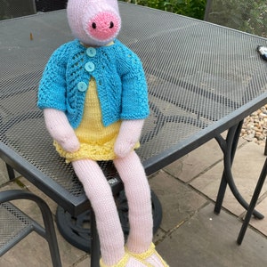 May include: A pink knitted pig wearing a blue cardigan and a yellow dress, sitting on a metal table with its legs dangling. The pig is wearing yellow shoes with blue trim.
