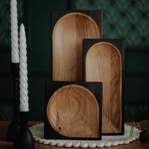 Könnte beinhalten: Ein Set aus drei Holzschalen mit dunklem Rahmen und hellem Holzinnenraum. Die Schalen sind in einer Reihe angeordnet, mit einer quadratischen Schale vorne und zwei rechteckigen Schalen dahinter. Zwei weiße Spiral-Kerzen in einem schwarzen Kerzenhalter.