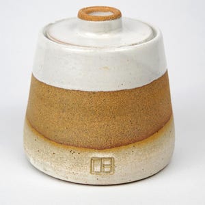 May include: A ceramic sugar bowl with a lid. The bowl is light beige at the base, transitioning to a tan middle section, and topped with a white band. The lid has a small, circular handle.