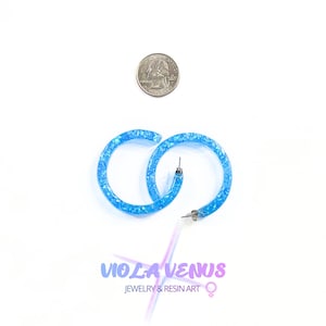 May include: A pair of bright blue resin hoop earrings with embedded glitter, shown next to a US quarter coin for scale. The text below reads VIOLA VENUS JEWELRY & RESIN ART with a female gender symbol. The earrings feature silver-toned metal posts.