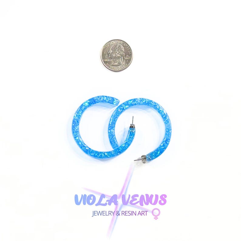May include: A pair of bright blue resin hoop earrings with embedded glitter, shown next to a US quarter coin for scale. The text below reads VIOLA VENUS JEWELRY & RESIN ART with a female gender symbol. The earrings feature silver-toned metal posts.