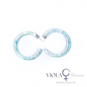 May include: A pair of clear resin hoop earrings featuring embedded iridescent blue, purple, and green flakes. The earrings have silver-toned metal posts. The text VIOLA Venus JEWELRY & RESIN ART is printed in the bottom right corner of the image.