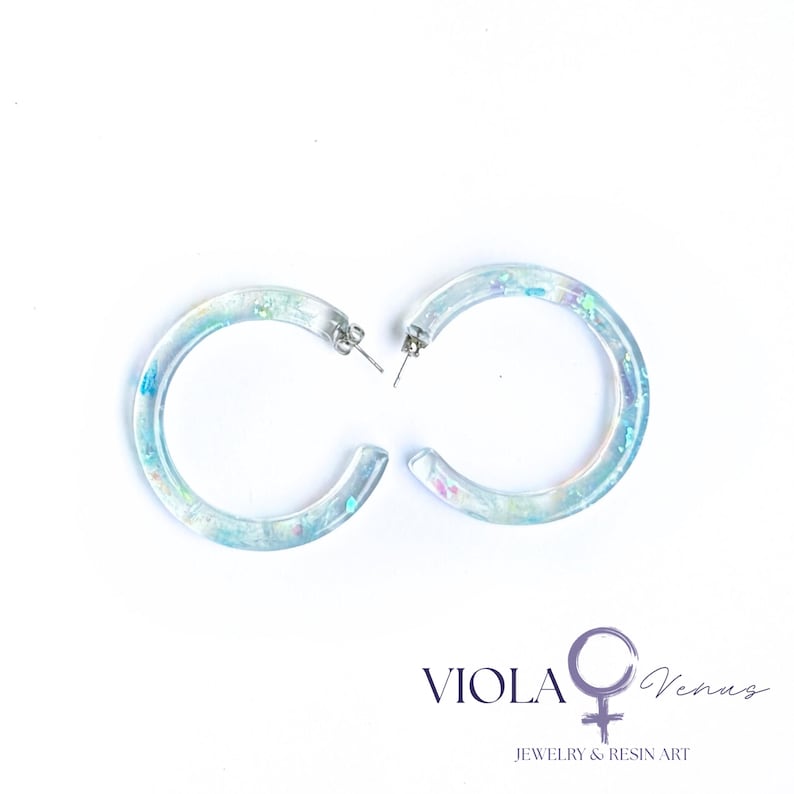 May include: A pair of clear resin hoop earrings featuring embedded iridescent blue, purple, and green flakes. The earrings have silver-toned metal posts. The text VIOLA Venus JEWELRY & RESIN ART is printed in the bottom right corner of the image.