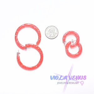 May include: Two pairs of coral-colored resin hoop earrings with embedded metallic flakes. The larger pair is positioned next to a US quarter coin for scale, while the smaller pair is placed to the right. Each earring features a silver-tone post.