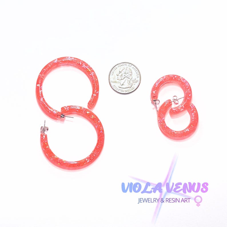 May include: Two pairs of coral-colored resin hoop earrings with embedded metallic flakes. The larger pair is positioned next to a US quarter coin for scale, while the smaller pair is placed to the right. Each earring features a silver-tone post.