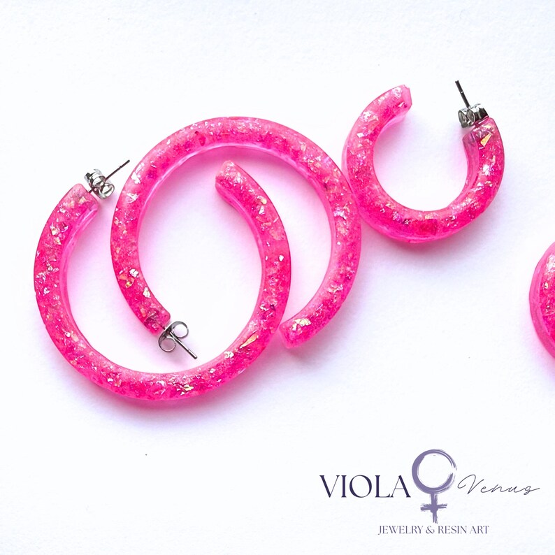 May include: Bright pink resin hoop earrings featuring embedded iridescent glitter flakes. The set includes two large hoops and one smaller hoop, each with silver-tone metal posts and backings. The text VIOLA Venus JEWELRY & RESIN ART is visible.
