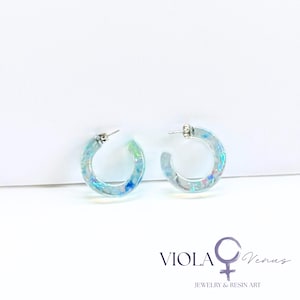 May include: A pair of clear resin hoop earrings featuring embedded iridescent blue and green flakes. The earrings have silver-toned metal posts. The text VIOLA Venus JEWELRY & RESIN ART is printed in the bottom right corner of the image.