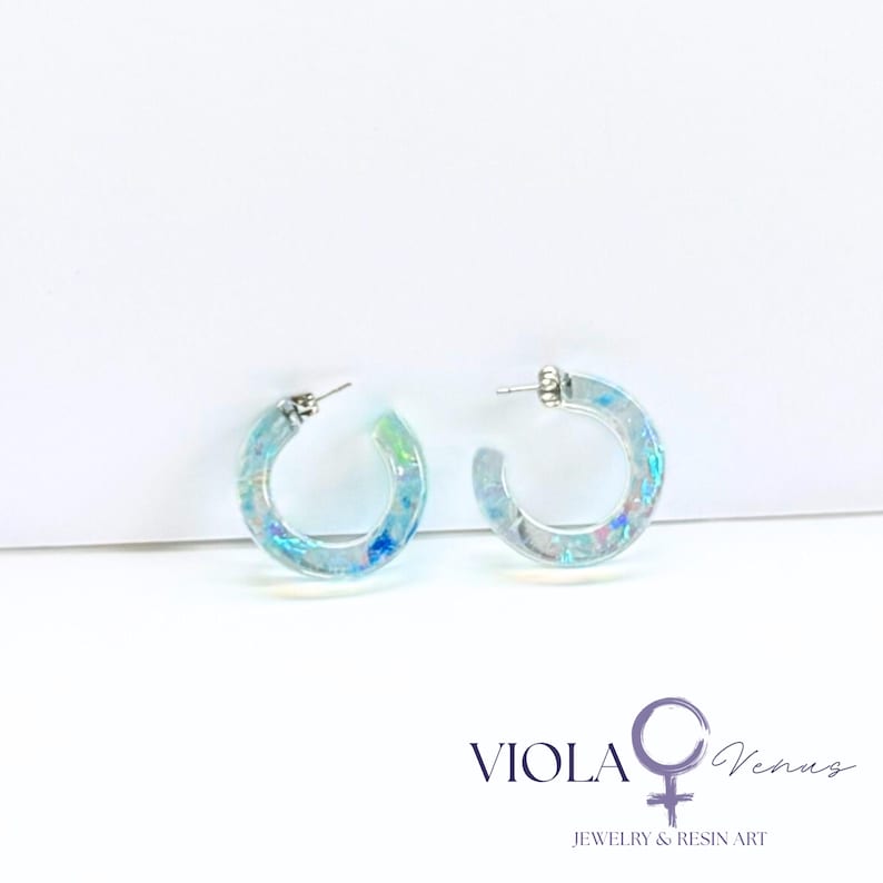 May include: A pair of clear resin hoop earrings featuring embedded iridescent blue and green flakes. The earrings have silver-toned metal posts. The text VIOLA Venus JEWELRY & RESIN ART is printed in the bottom right corner of the image.