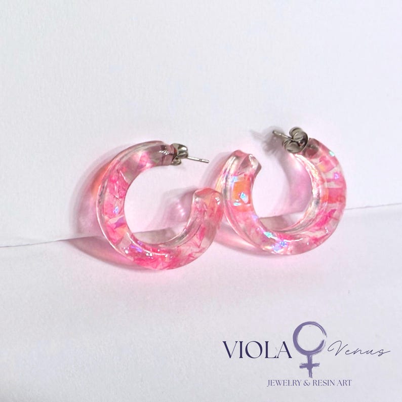 May include: A pair of hoop earrings crafted from clear resin, featuring embedded pink and iridescent flakes that catch the light. The earrings have silver-toned metal posts and backings. The text VIOLA Venus JEWELRY & RESIN ART is visible.