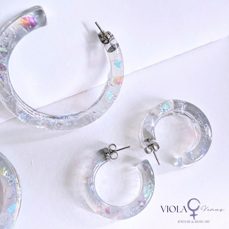 May include: Clear resin hoop earrings featuring embedded iridescent flakes that reflect light in various colors. The set includes one large hoop and two smaller hoops, each equipped with silver-toned metal posts. Text reads VIOLA Venus JEWELRY & RESIN ART.