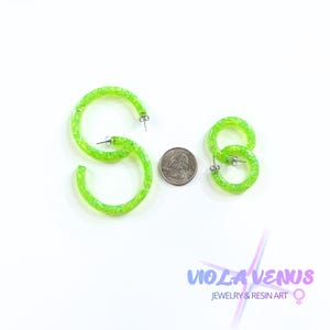 May include: Two pairs of neon green hoop earrings with embedded iridescent glitter flakes. The larger pair is positioned on the left, and the smaller pair is on the right. A United States quarter coin is placed between the two sets for scale.